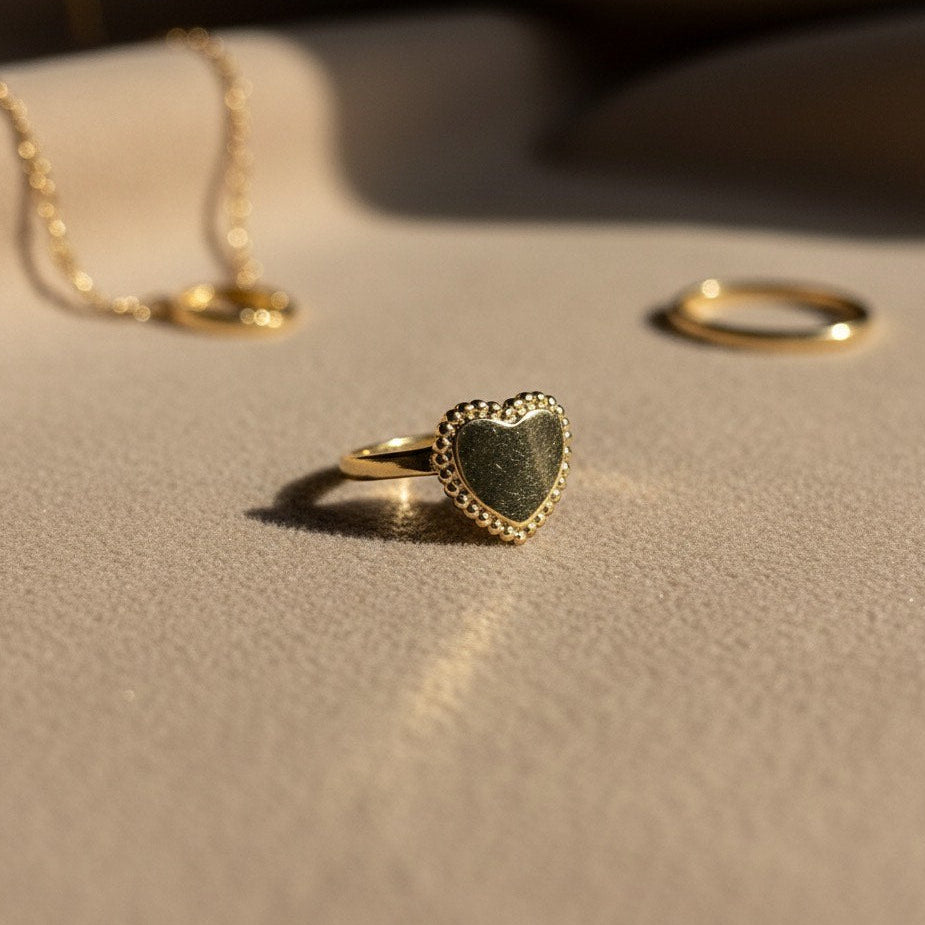 Heart-shaped gold ring on a beige surface with a necklace and another ring in the background.