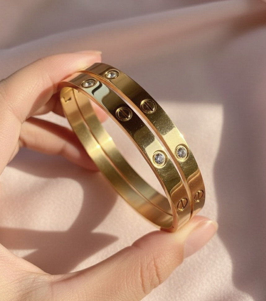 Gold bangles with embedded stones held by a hand against a soft pink background