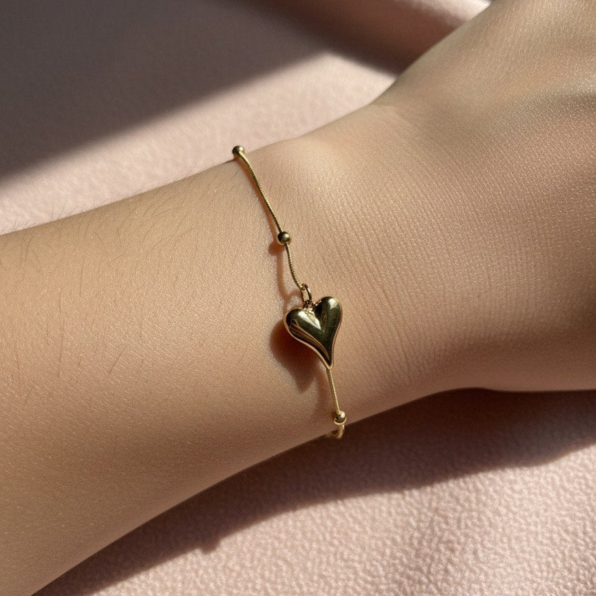 Gold bracelet with a heart charm on a wrist against a soft pink background