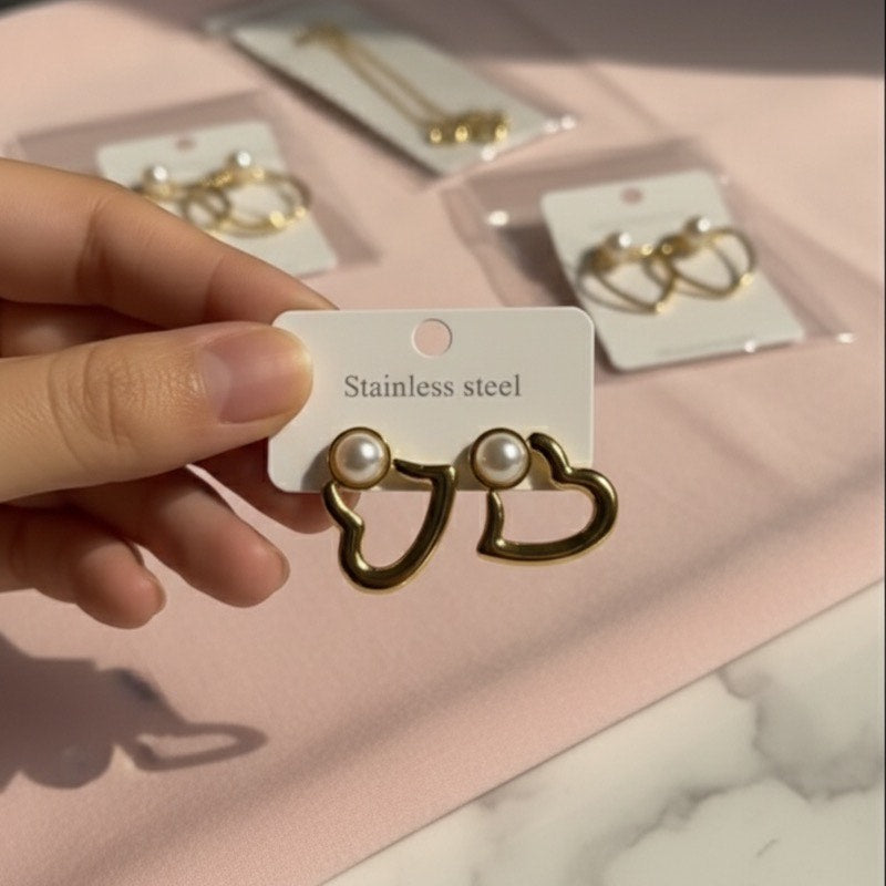 Gold heart-shaped earrings with pearls held by a hand on a pink surface with more earrings in the background.